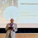 Piiskopirüüs vaimulik seisab mikrofoni ja tahvelarvutiga laval, tema taga ekraanil Eesti Kalmistute Liidu infopäeva tiitelslaid.